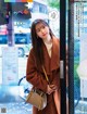 A woman in a brown coat is standing in front of a window.